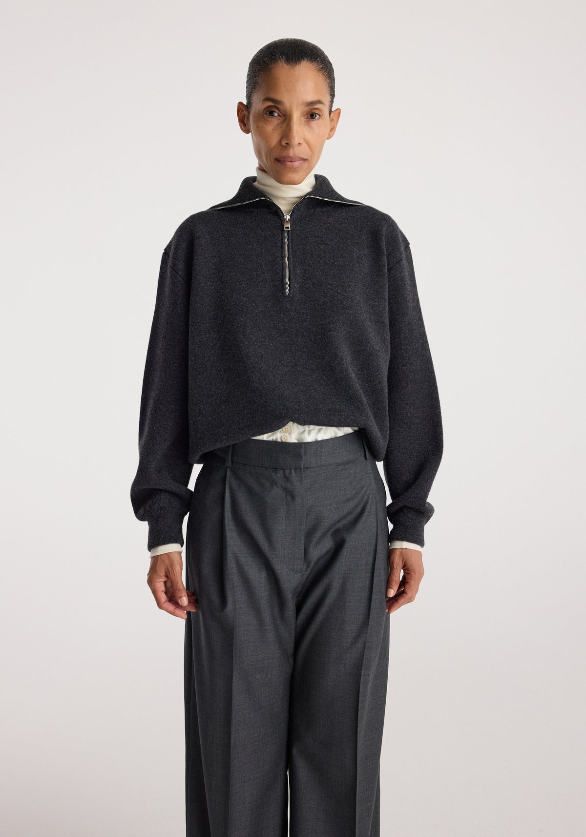 Woman wearing a charcoal Two-way zipped turtleneck | charcoal with long sleeves over light inner layer, paired with high-waisted dark gray tailored pants against plain background.