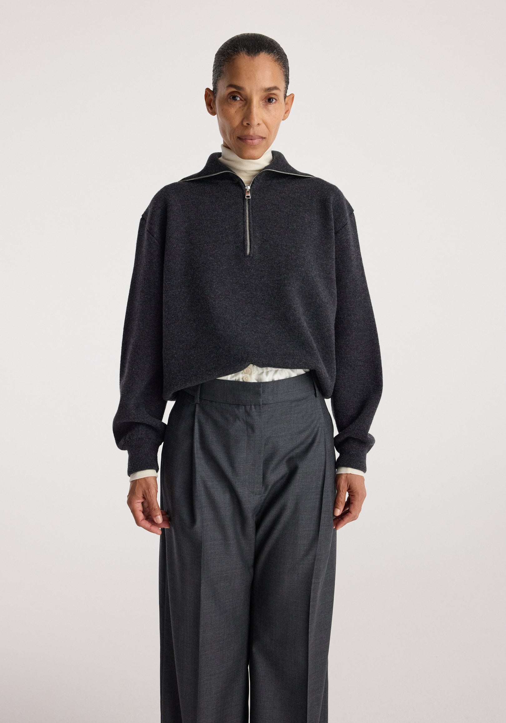 Woman wearing a charcoal Two-way zipped turtleneck | charcoal with long sleeves over light inner layer, paired with high-waisted dark gray tailored pants against plain background.