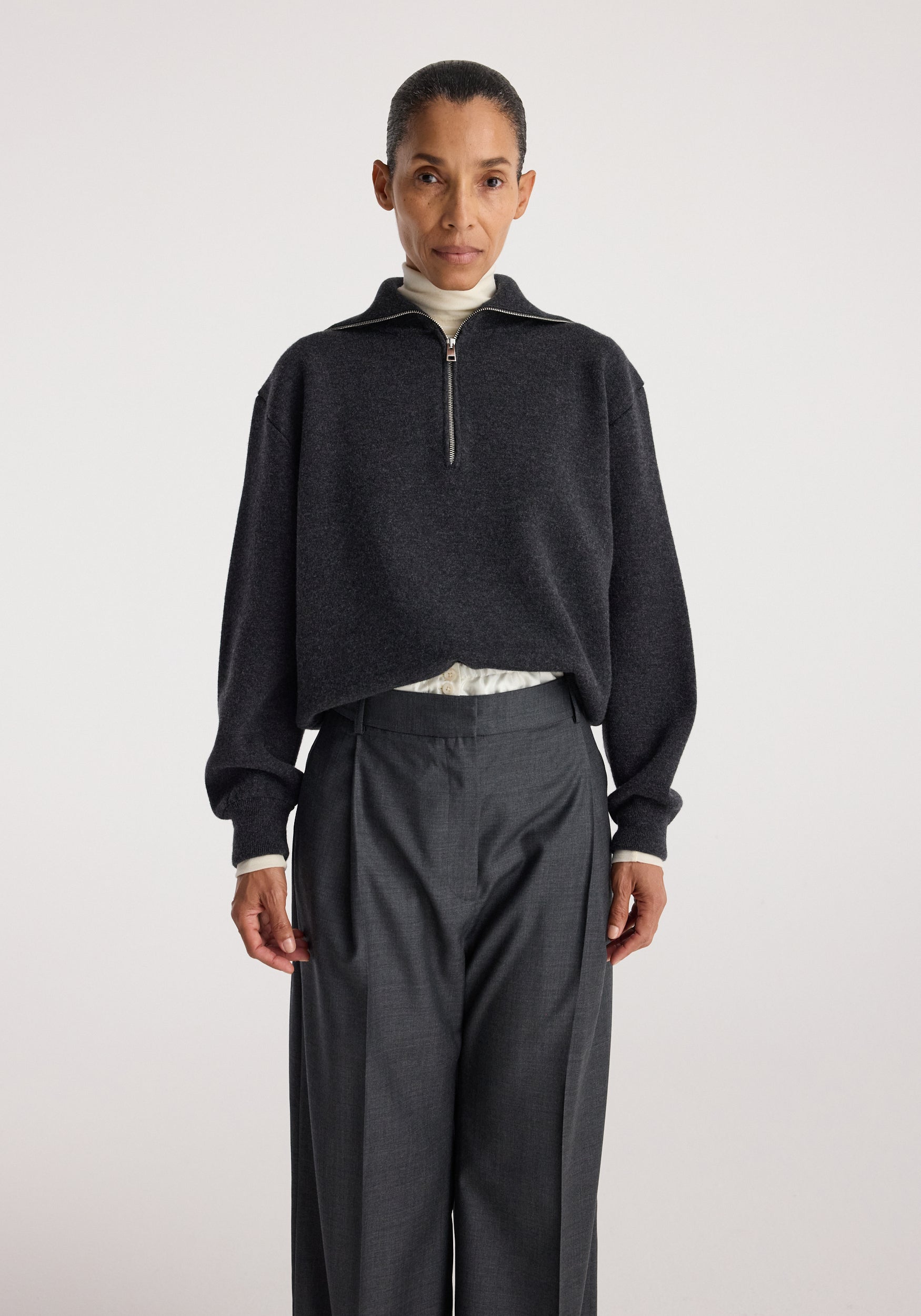 Woman wearing a charcoal Two-way zipped turtleneck | charcoal with long sleeves over light inner layer, paired with high-waisted dark gray tailored pants against plain background.