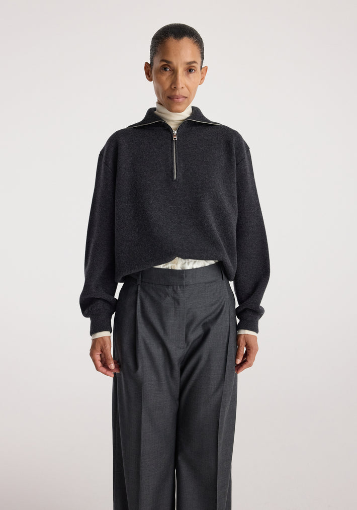 Woman wearing a charcoal Two-way zipped turtleneck | charcoal with long sleeves over light inner layer, paired with high-waisted dark gray tailored pants against plain background.