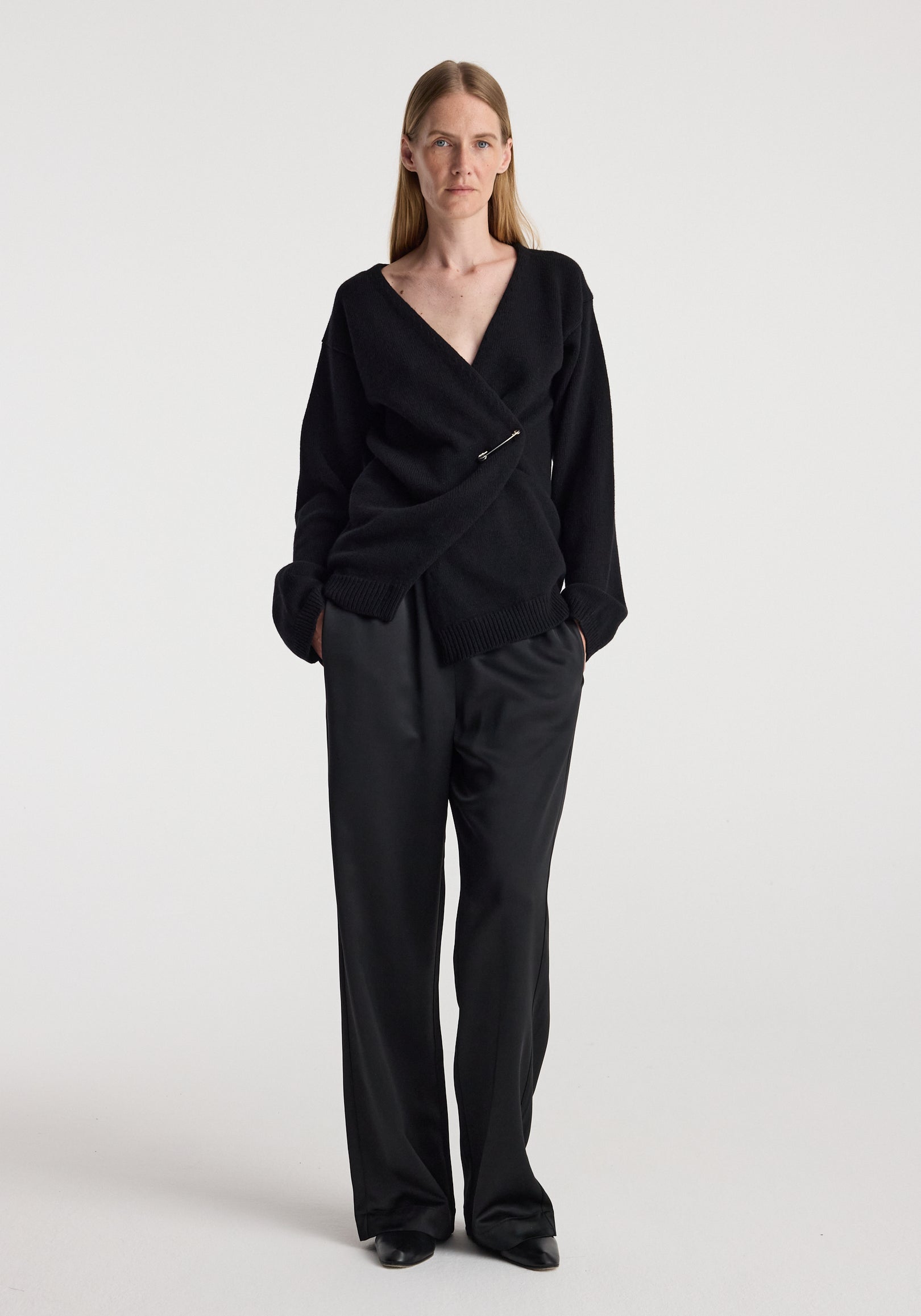 Woman wearing a black off-shoulder wrap cardigan | black with long sleeves and a pin closure, styled with loose black pants against a plain light background.