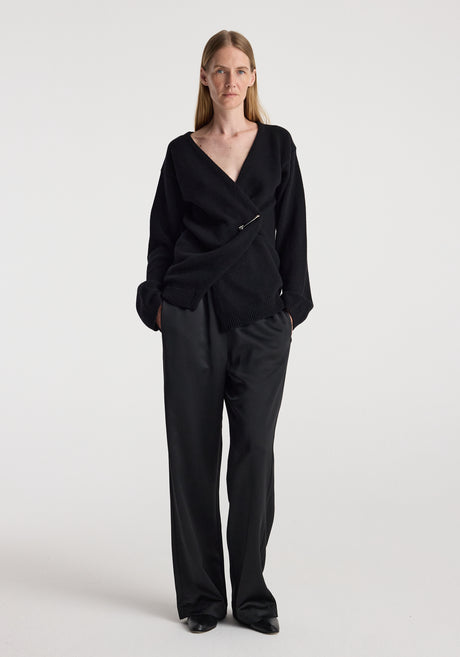 Woman wearing a black off-shoulder wrap cardigan | black with long sleeves and a pin closure, styled with loose black pants against a plain light background.