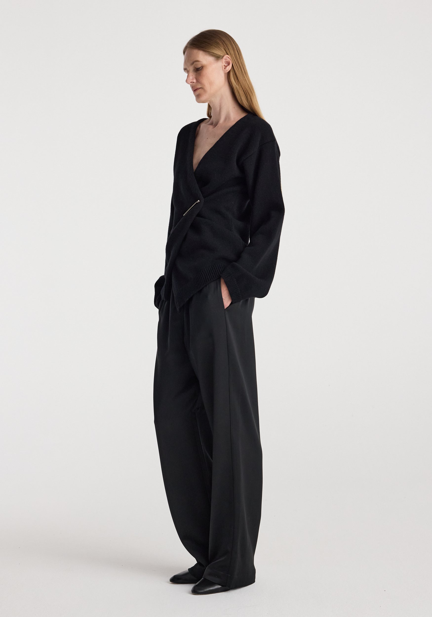 Woman wearing an elegant Off-shoulder wrap cardigan | black paired with loose black trousers, standing against a plain white background in a relaxed pose.
