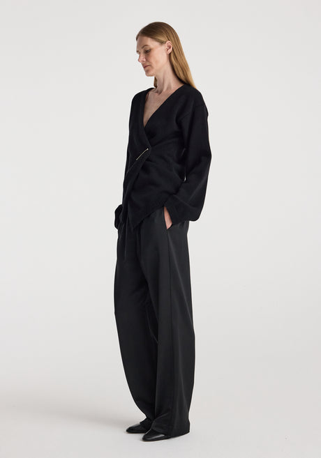 Woman wearing a black off-shoulder wrap cardigan | black with long sleeves and a pin closure, styled with loose black pants against a plain light background.