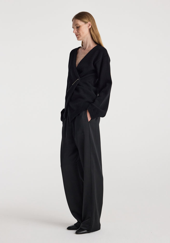 Woman wearing a black off-shoulder wrap cardigan | black with long sleeves and a pin closure, styled with loose black pants against a plain light background.