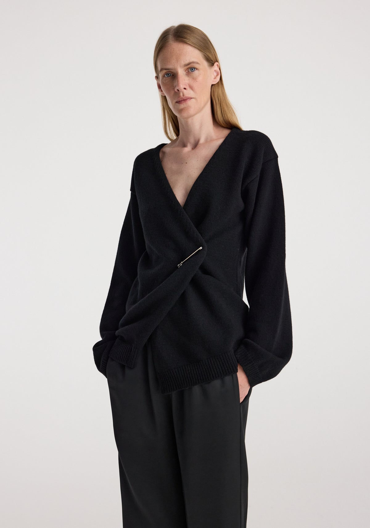 Woman wearing an off-shoulder wrap cardigan | black with a deep V-neck and long sleeves, styled with black pants against a plain background.