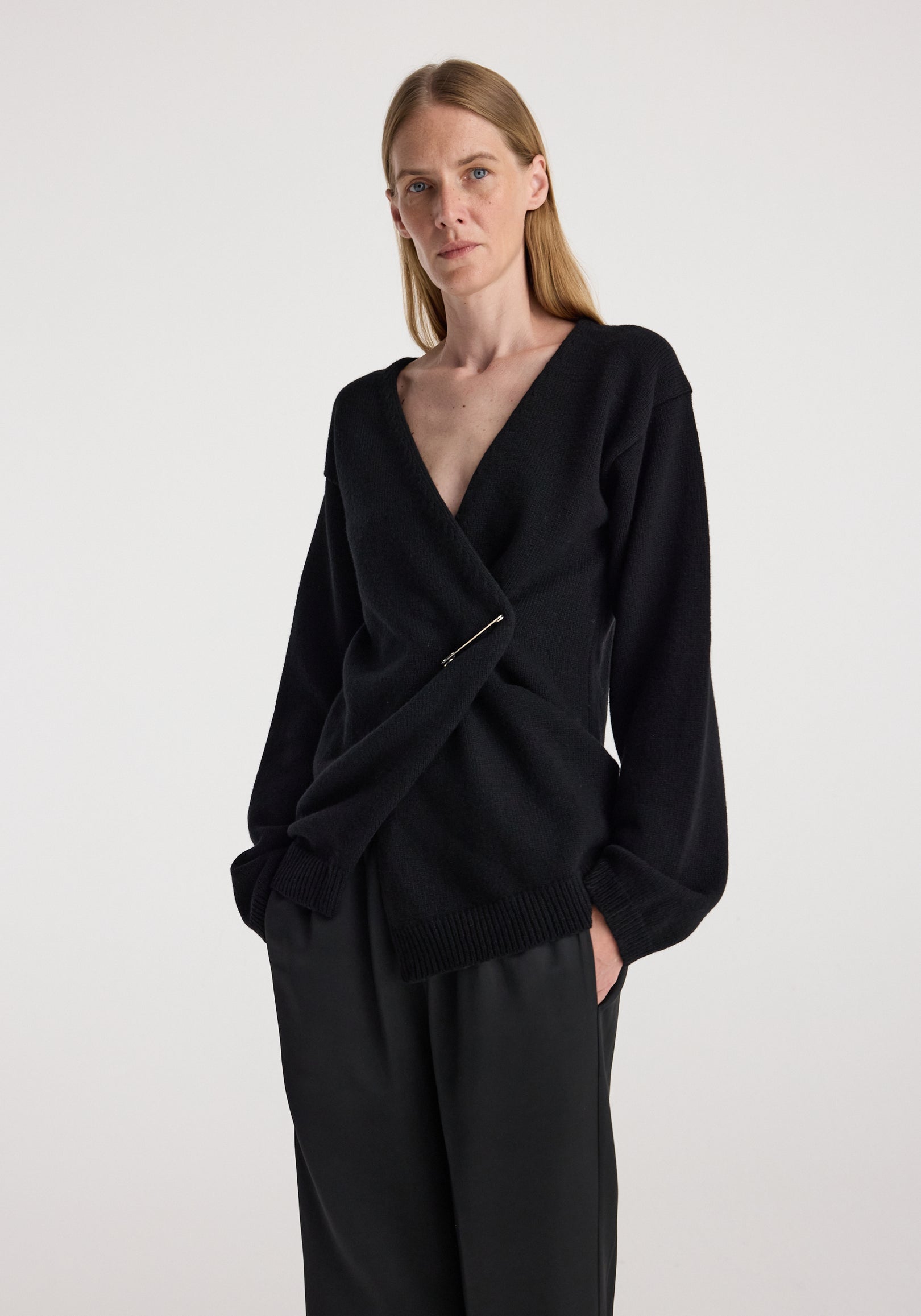Woman wearing an off-shoulder wrap cardigan | black with a deep V-neck and long sleeves, styled with black pants against a plain background.