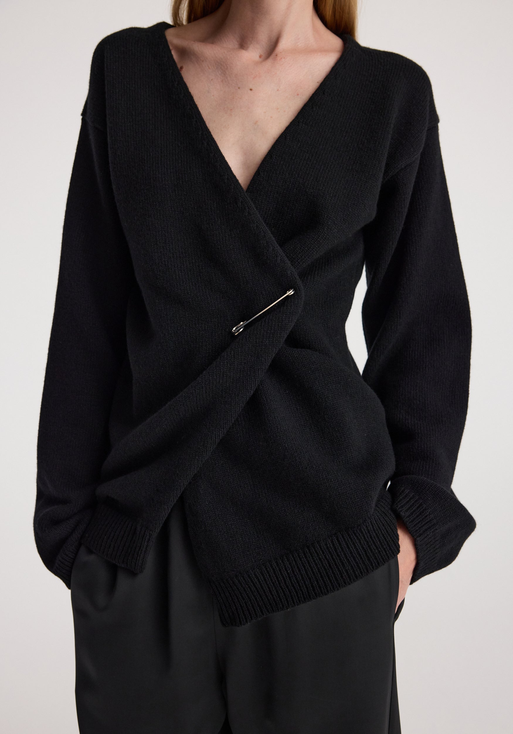 Woman wearing a black off-shoulder wrap cardigan | black with long sleeves and a safety pin fastening on the front, paired with black pants.