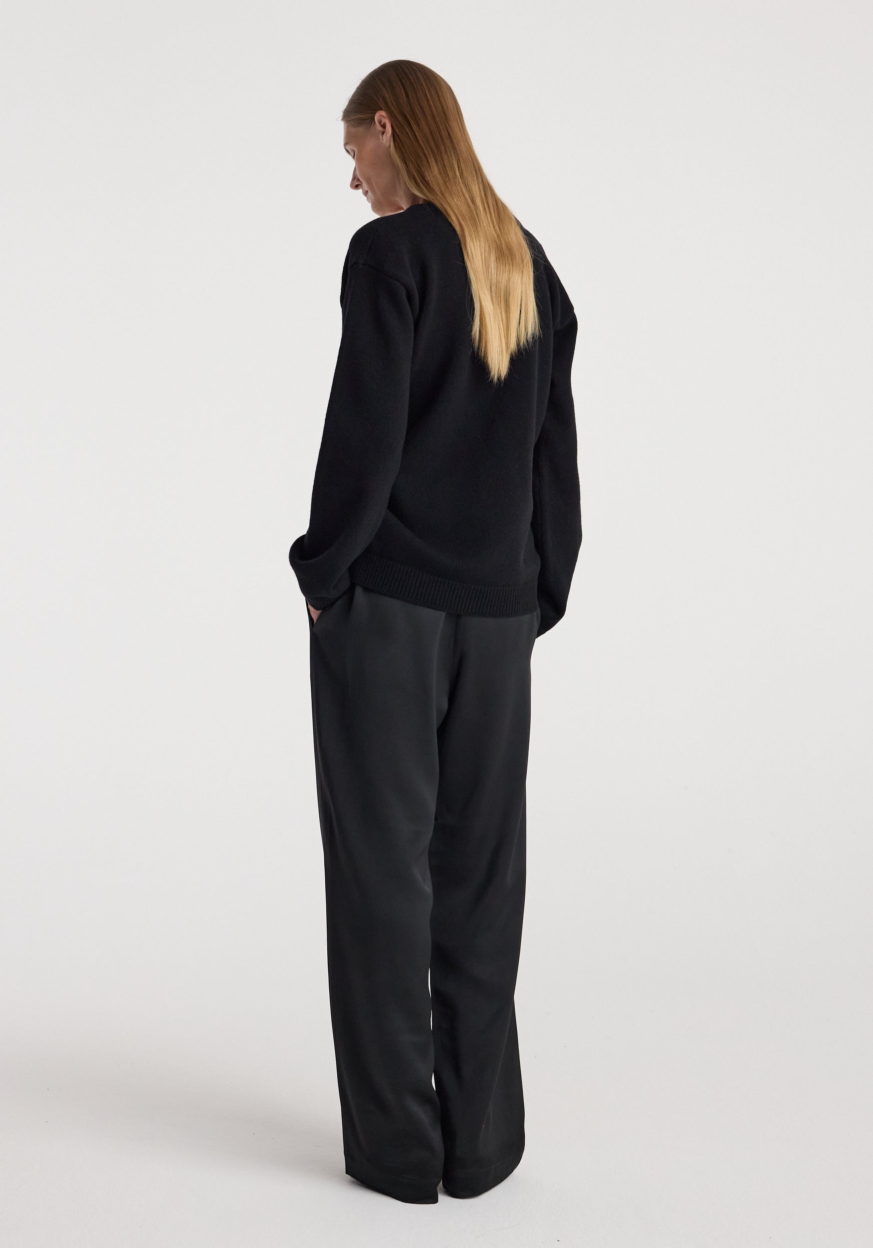 Woman standing with hands in pockets wearing an off-shoulder wrap cardigan | black from the back, paired with loose black pants against a plain background.