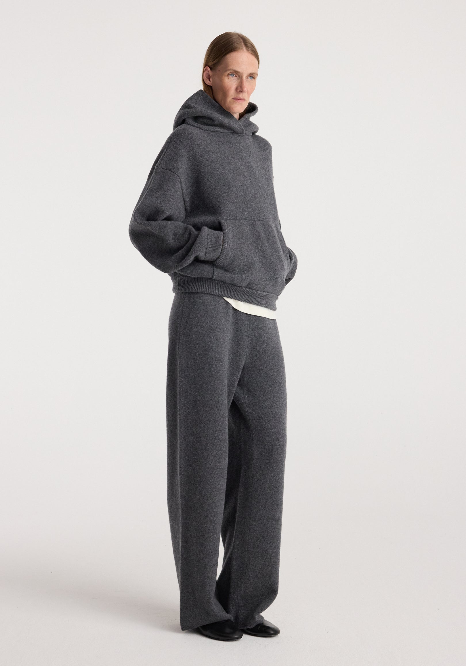 Woman wearing a comfortable and casual Hooded kangeroo jumper | dark grey melange with matching loose pants, standing against a plain background.