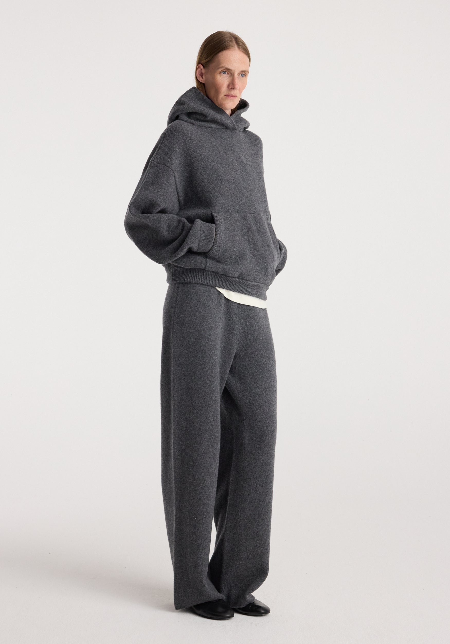 Woman wearing a comfortable and casual Hooded kangeroo jumper | dark grey melange with matching loose pants, standing against a plain background.