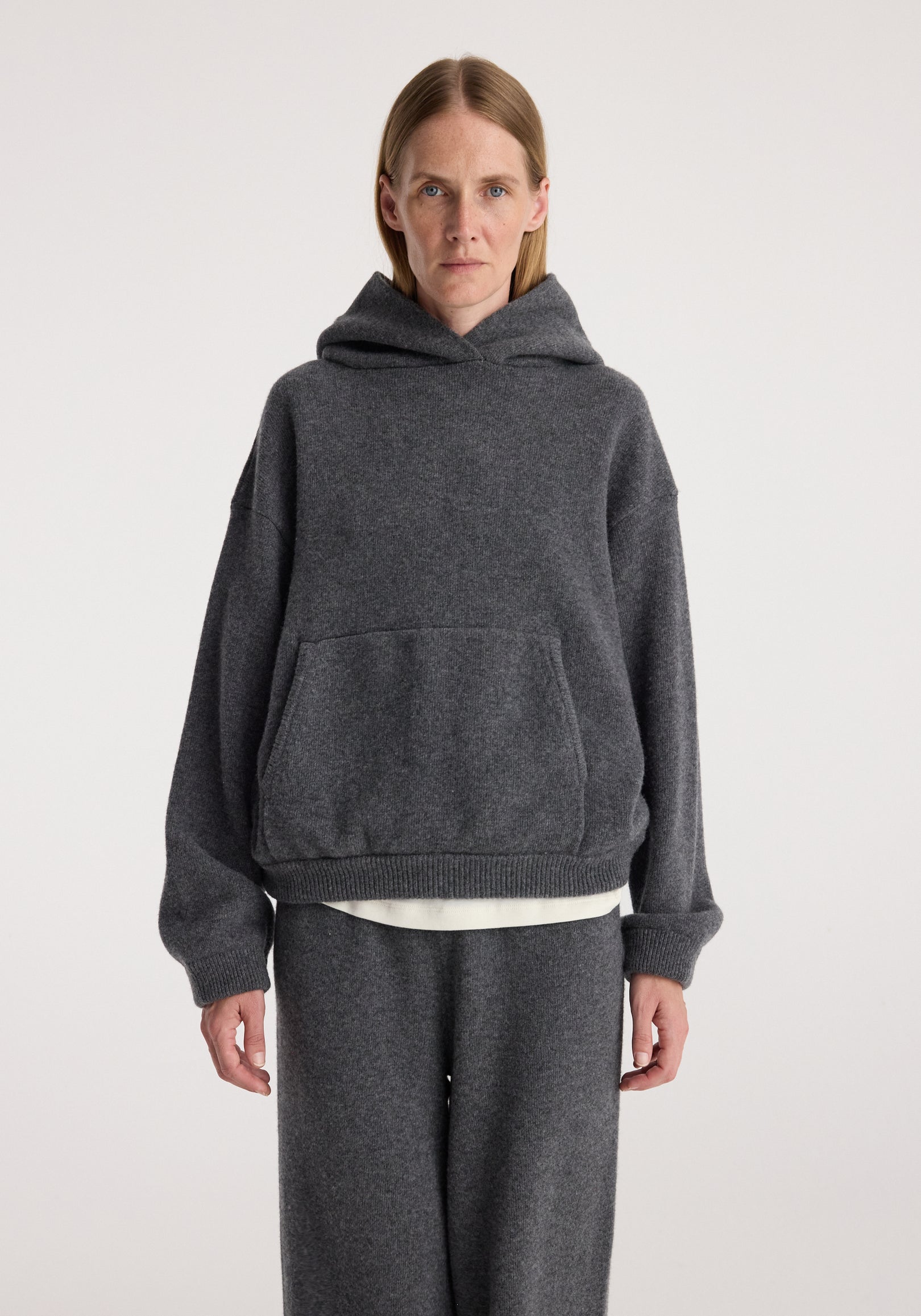 Woman wearing a dark grey melange hooded kangeroo jumper with a front pocket and relaxed fit, paired with matching dark grey pants against a plain background.