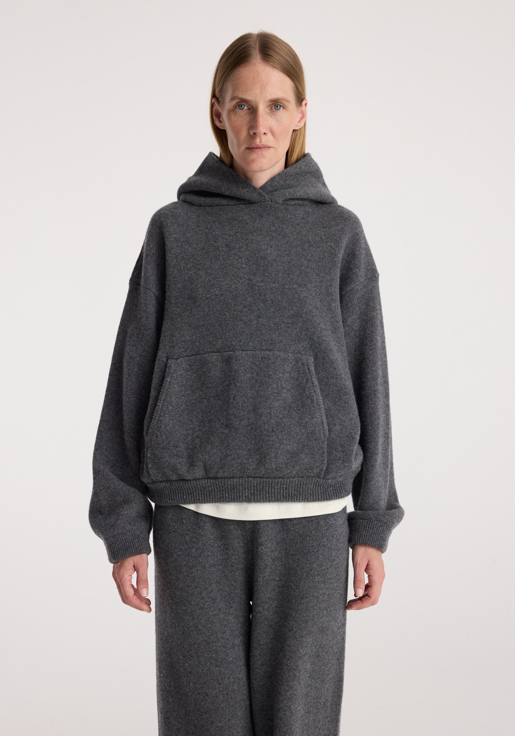 Woman wearing a dark grey melange hooded kangeroo jumper with a front pocket and relaxed fit, paired with matching dark grey pants against a plain background.
