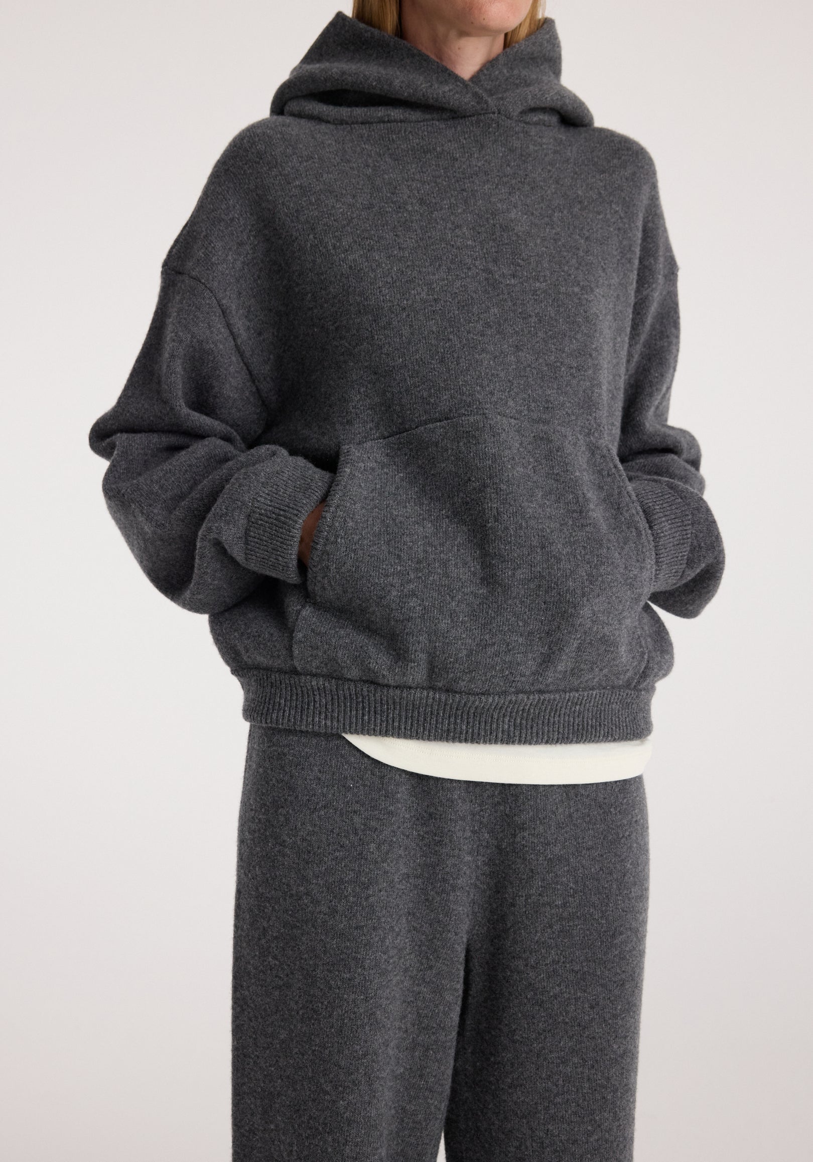 Person wearing a dark grey melange hooded kangeroo jumper with hands in front pocket, paired with matching dark grey pants and a white shirt peeking out underneath.