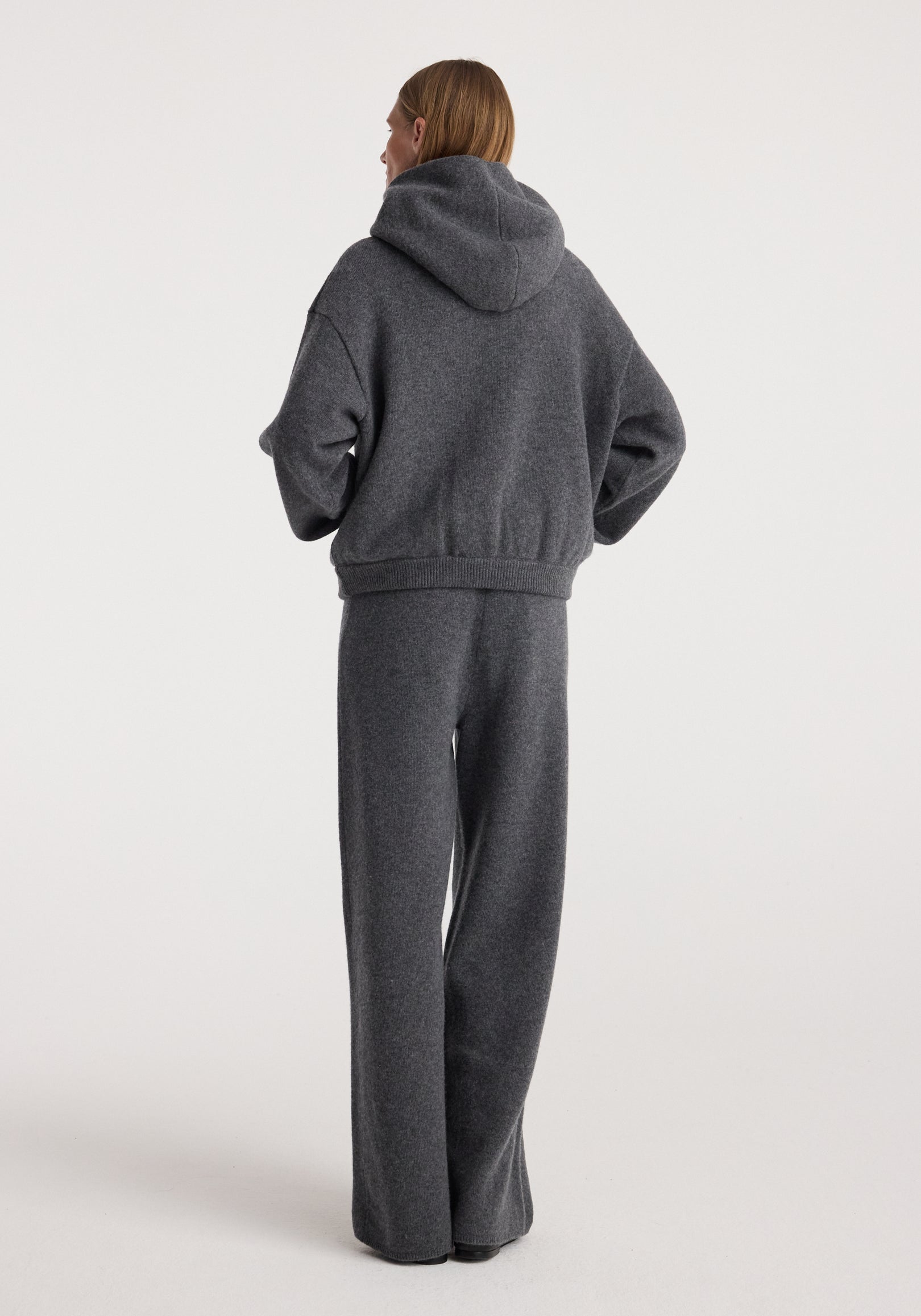 Back view of a person wearing a hooded kangeroo jumper | dark grey melange with matching loose-fitting pants against a plain background.
