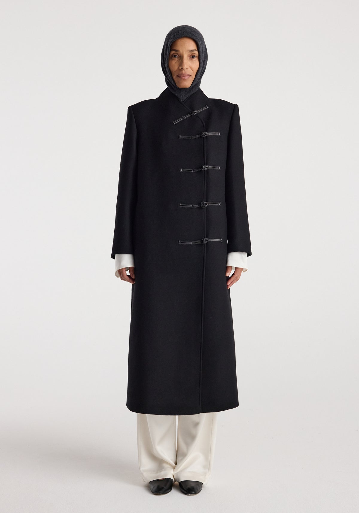 Woman wearing a black long coat with toggle closures and a Balaclava | anthracite melange hood, standing against a plain light background.