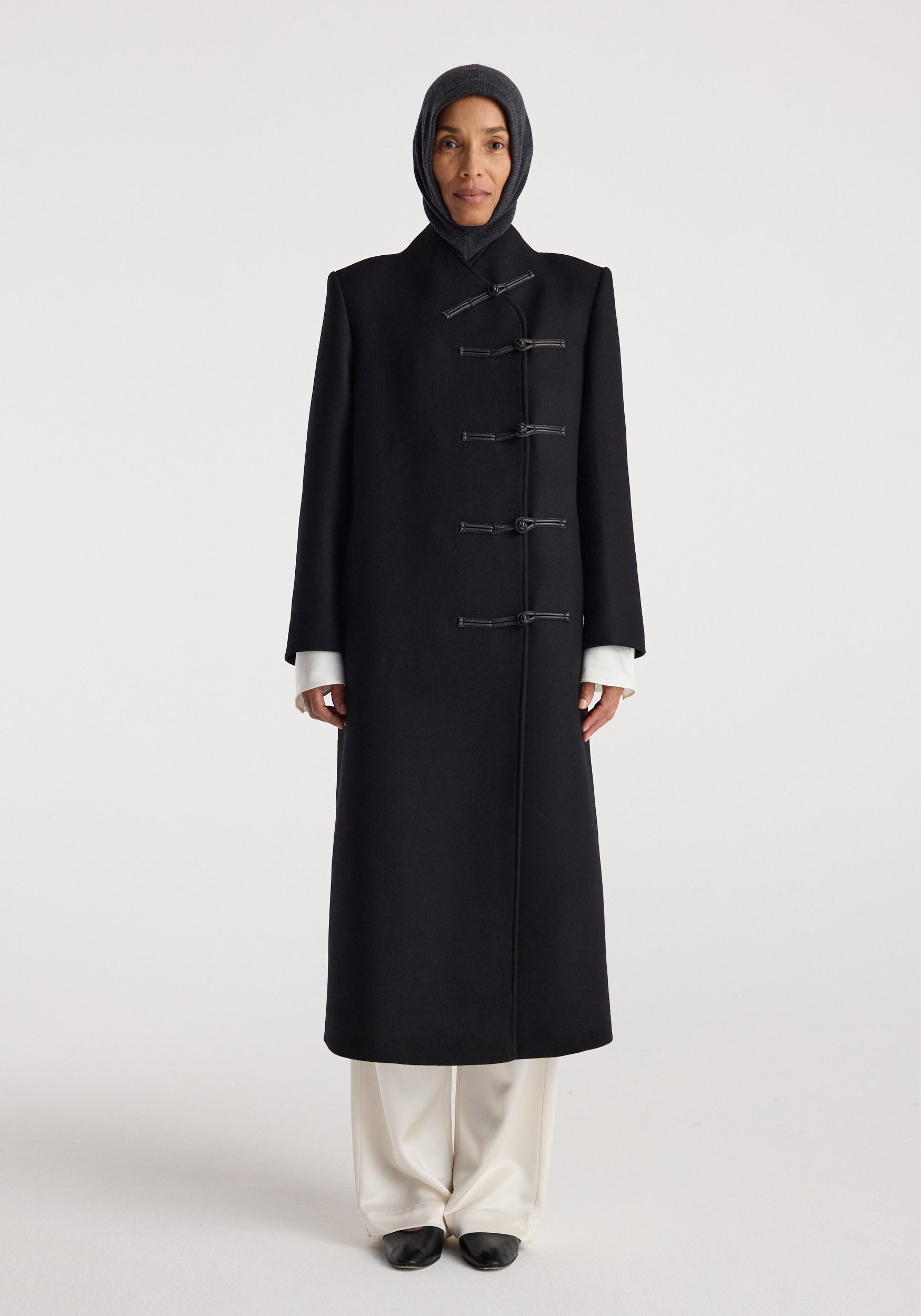 Woman wearing a black long coat with toggle closures and a Balaclava | anthracite melange hood, standing against a plain light background.