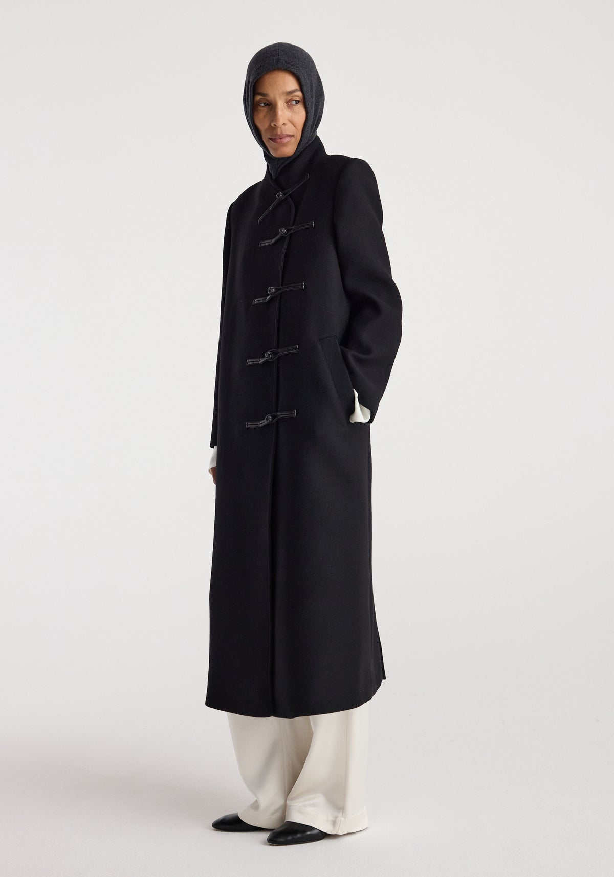 Woman wearing a dark grey knitted Balaclava | anthracite melange covering the head and neck, paired with a long black coat and white pants against a plain background.