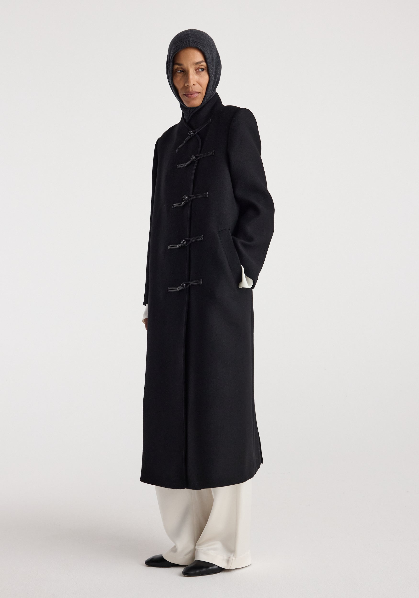 Woman wearing a dark grey knitted Balaclava | anthracite melange covering the head and neck, paired with a long black coat and white pants against a plain background.