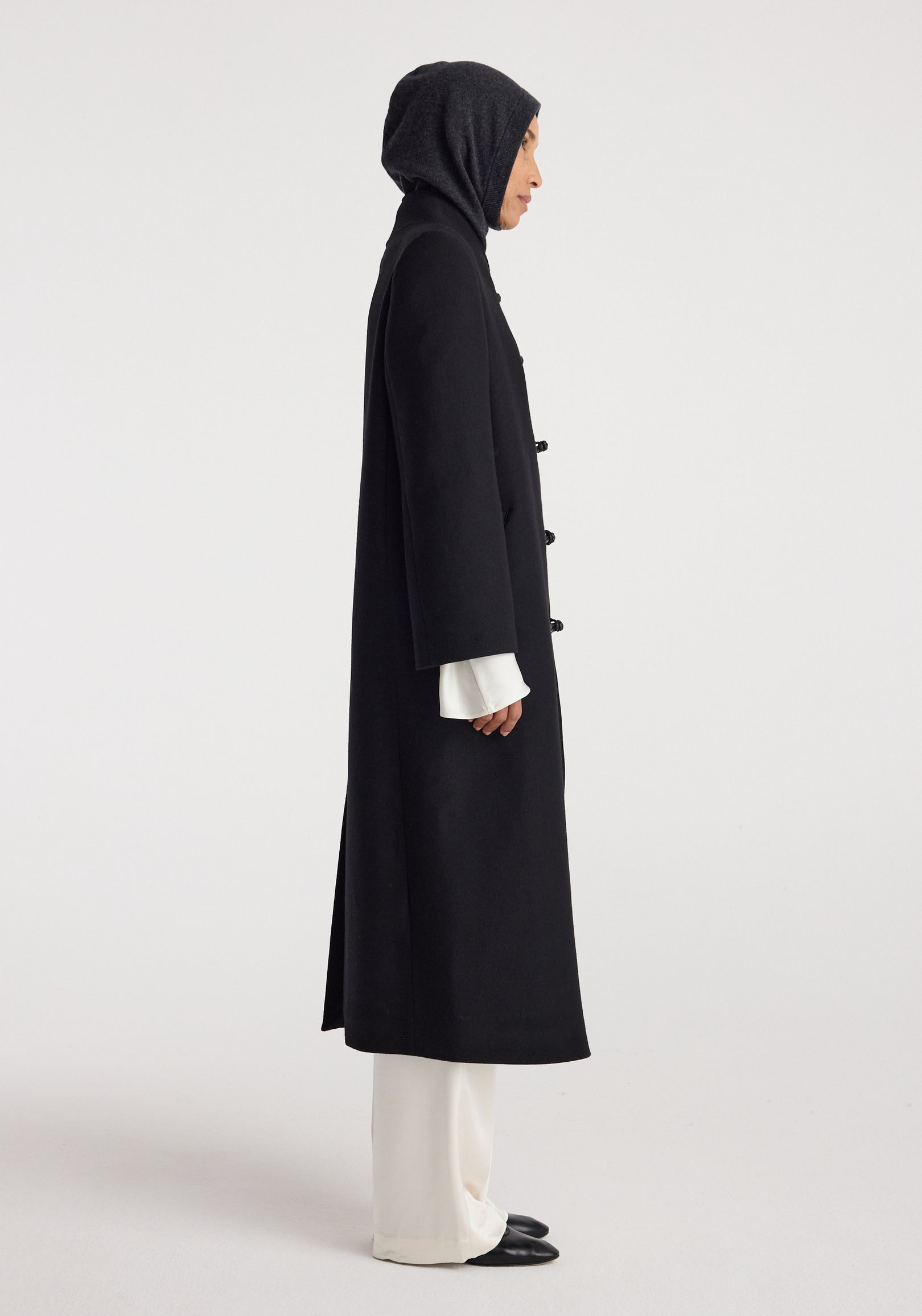 Woman wearing a dark hood over her head paired with a long black coat, white wide-legged pants, and black shoes, highlighting the Balaclava | anthracite melange.