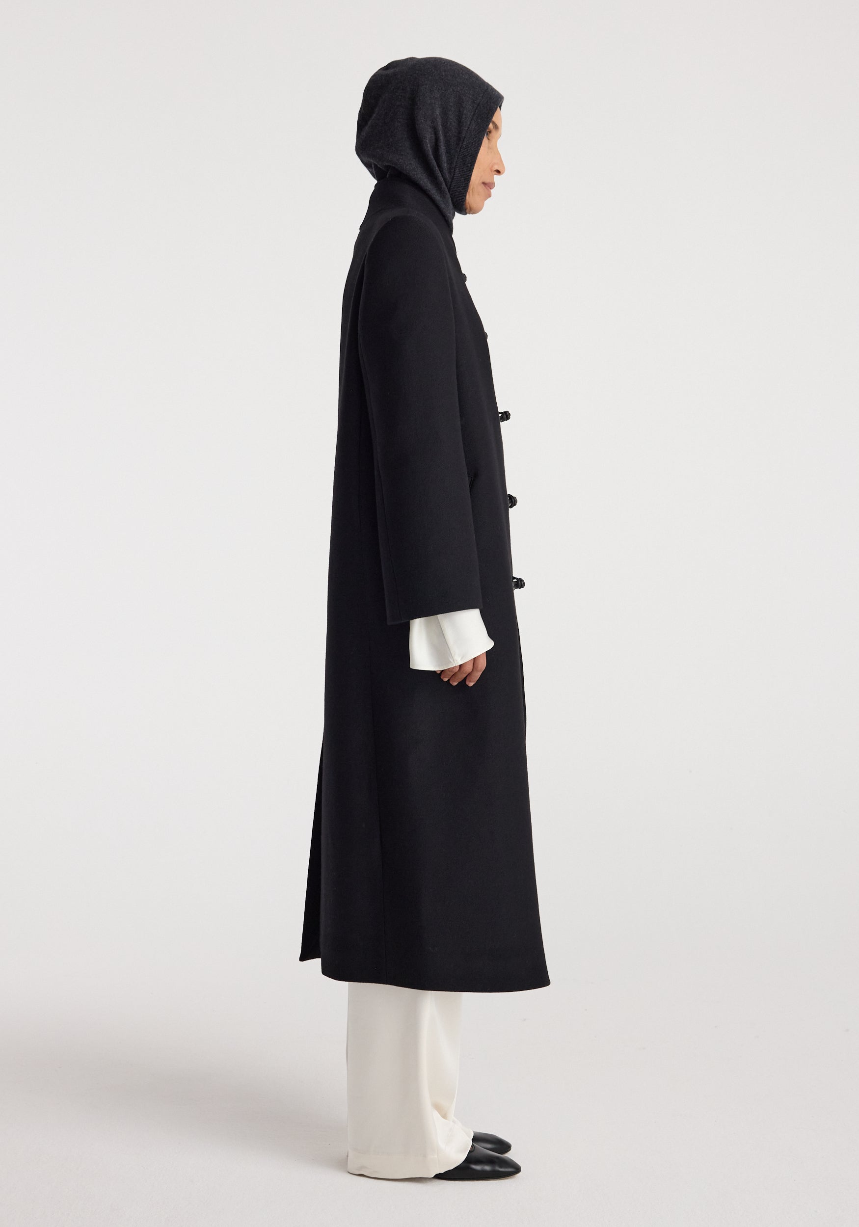 Woman wearing a dark hood over her head paired with a long black coat, white wide-legged pants, and black shoes, highlighting the Balaclava | anthracite melange.