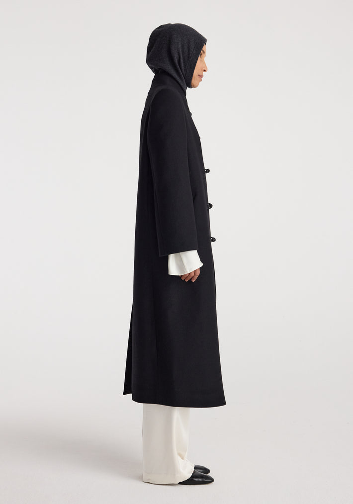 Woman wearing a dark hood over her head paired with a long black coat, white wide-legged pants, and black shoes, highlighting the Balaclava | anthracite melange.