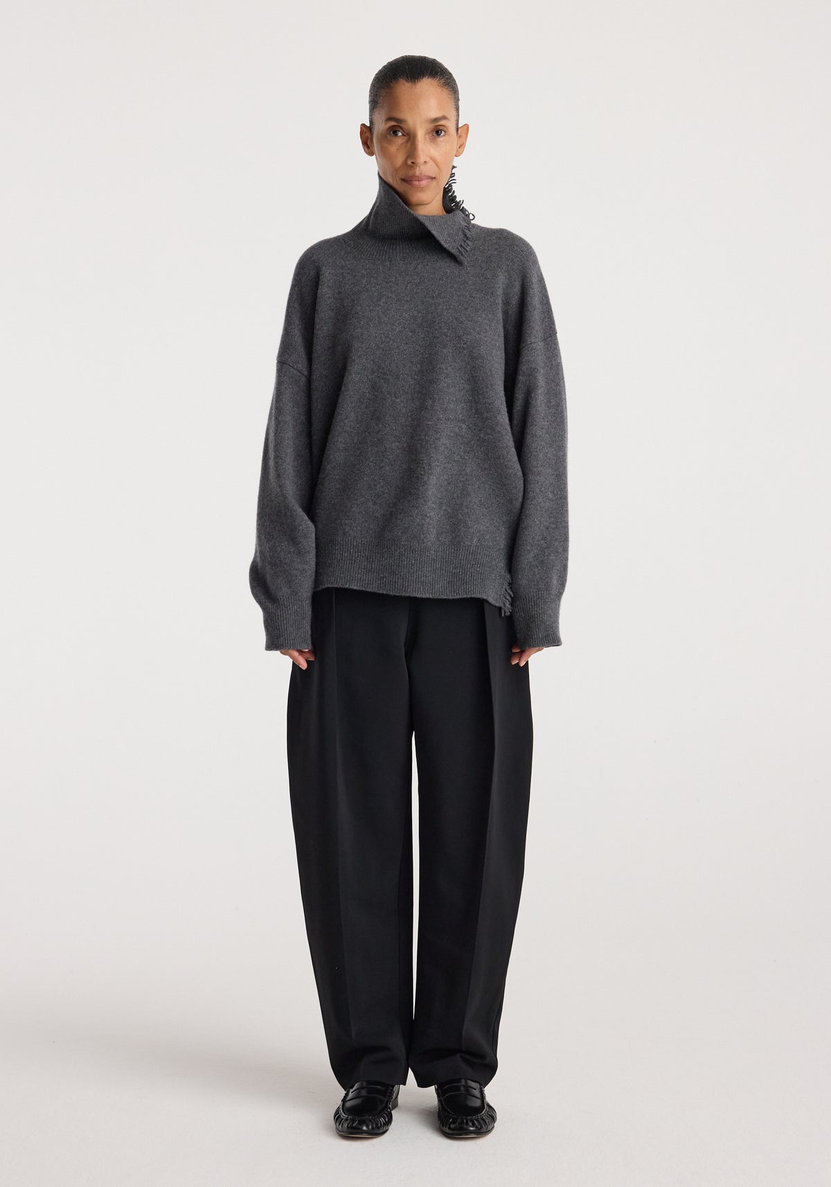Model wearing a loose-fitting dark grey melange Turtleneck with raw details paired with wide-leg black trousers and black shoes against a plain white background.
