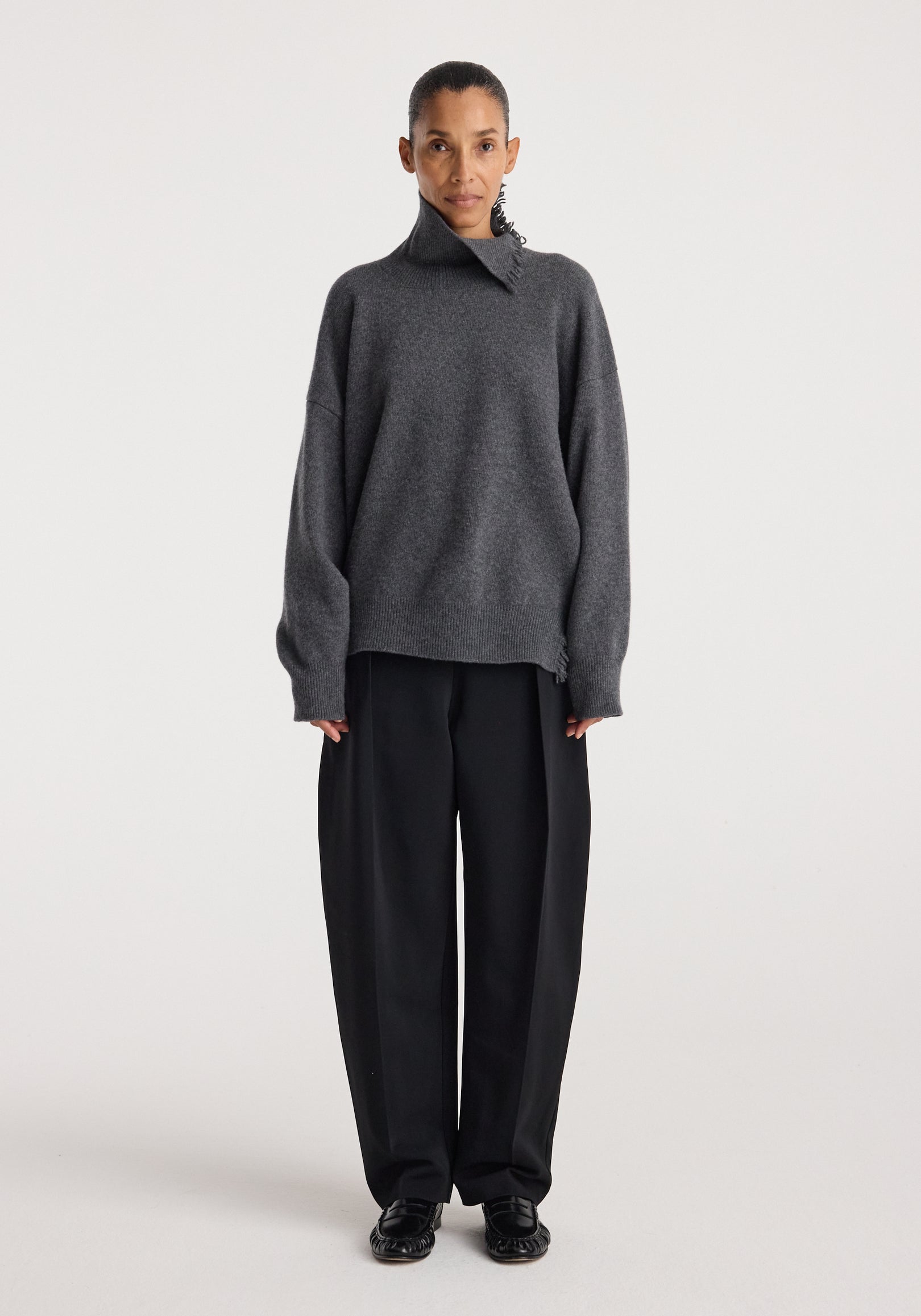 Model wearing a loose-fitting dark grey melange Turtleneck with raw details paired with wide-leg black trousers and black shoes against a plain white background.