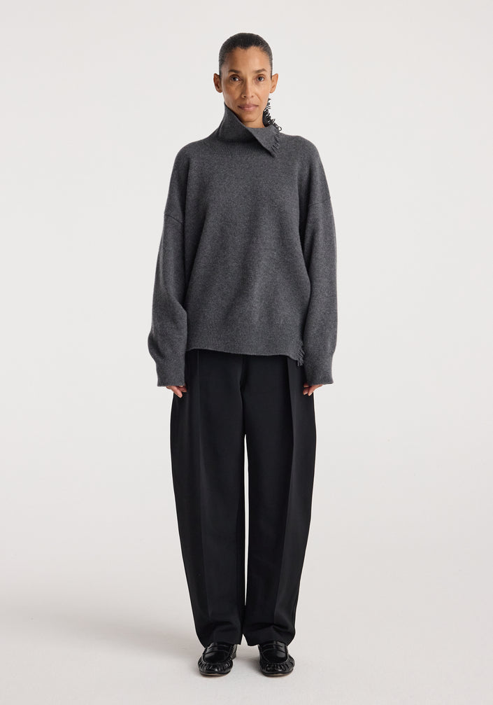 Model wearing a loose-fitting dark grey melange Turtleneck with raw details paired with wide-leg black trousers and black shoes against a plain white background.