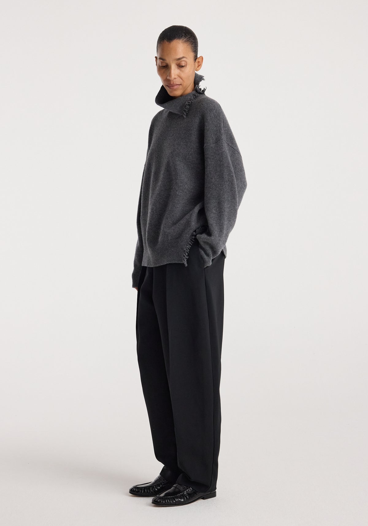 Woman wearing a loose-fitting Turtleneck with raw details | dark grey melange, paired with wide-leg black trousers and black shoes, standing against a plain white background.