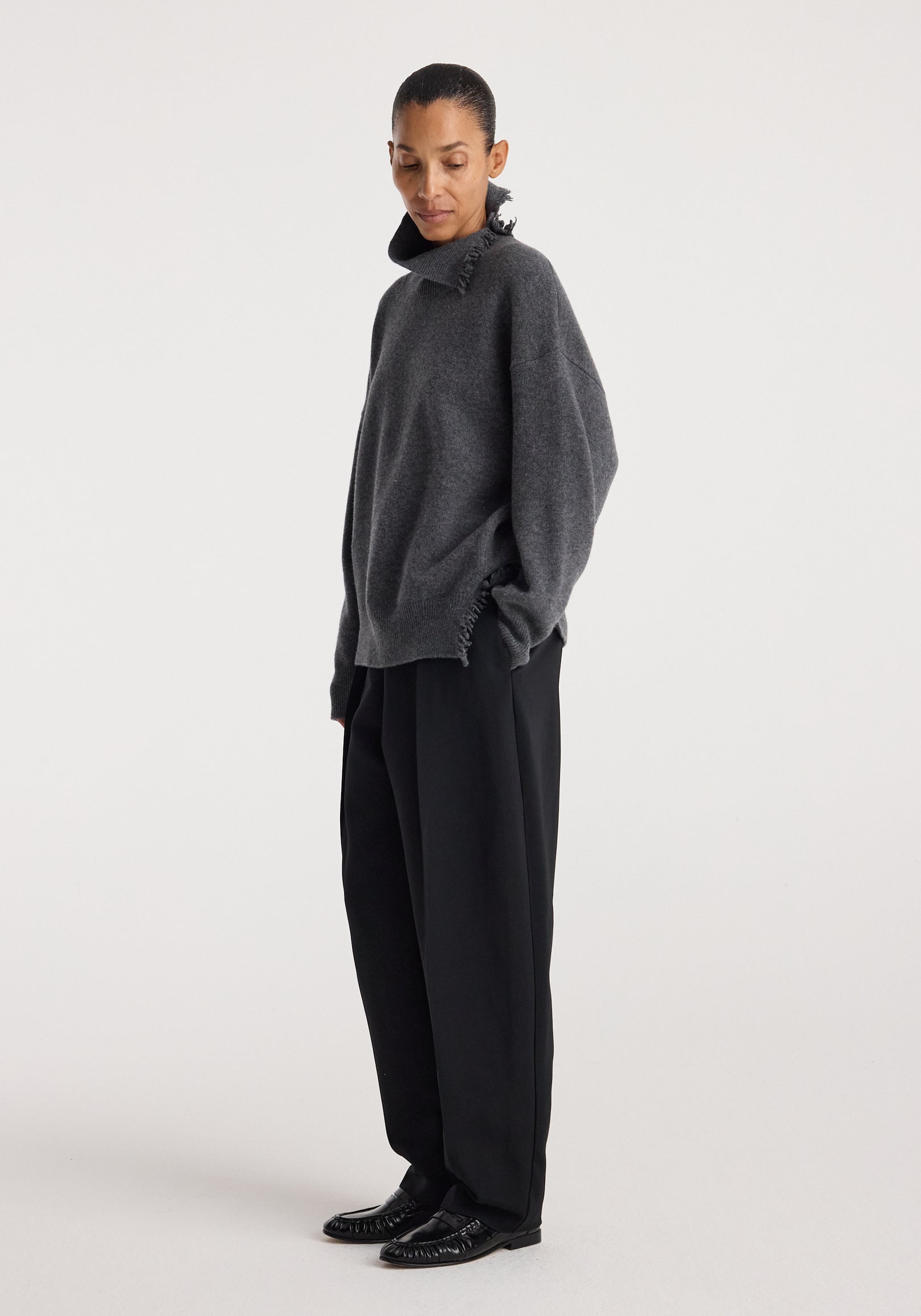 Woman wearing a loose-fitting Turtleneck with raw details | dark grey melange, paired with wide-leg black trousers and black shoes, standing against a plain white background.