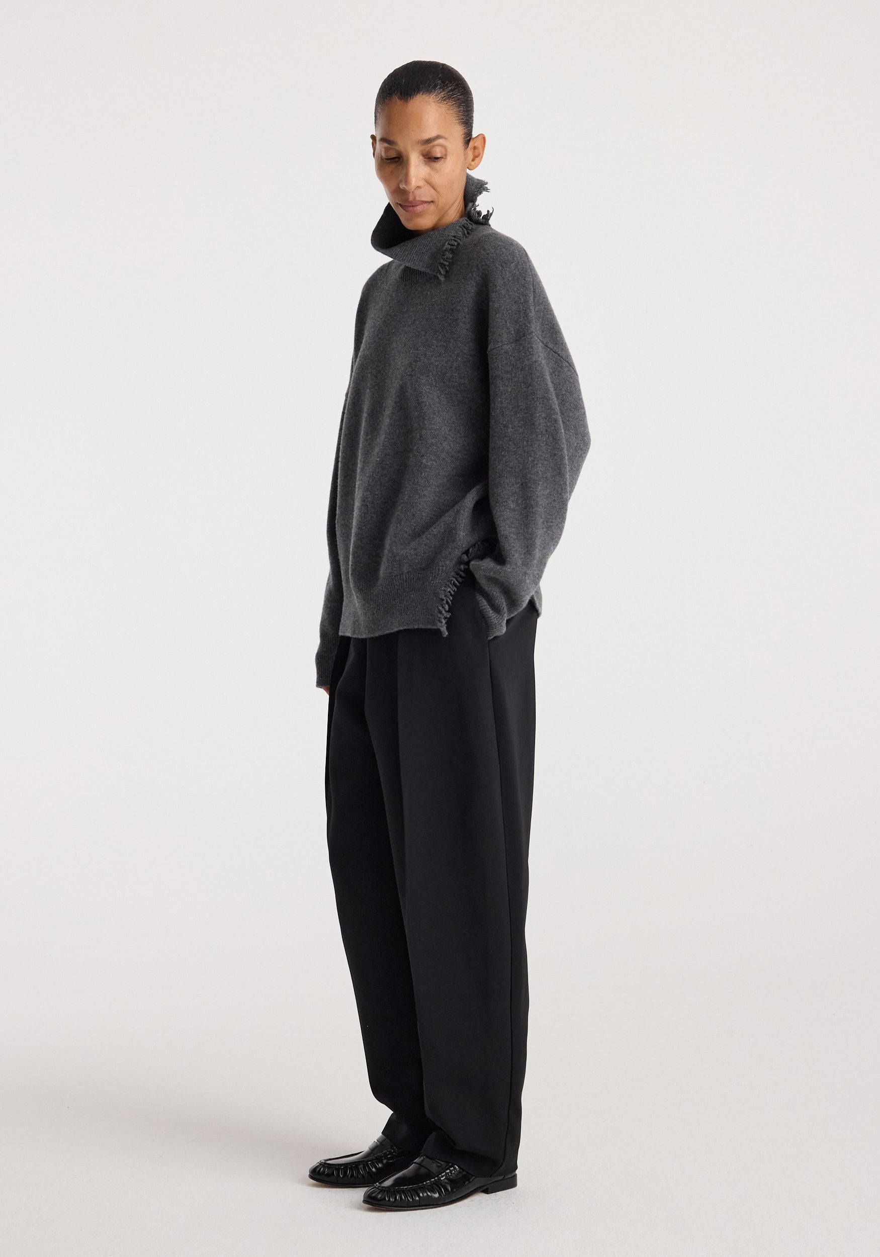 Woman wearing a loose-fitting Turtleneck with raw details | dark grey melange, paired with wide-leg black trousers and black shoes, standing against a plain white background.