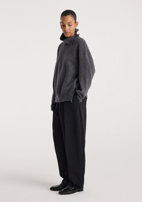 Model wearing a loose-fitting dark grey melange Turtleneck with raw details paired with wide-leg black trousers and black shoes against a plain white background.