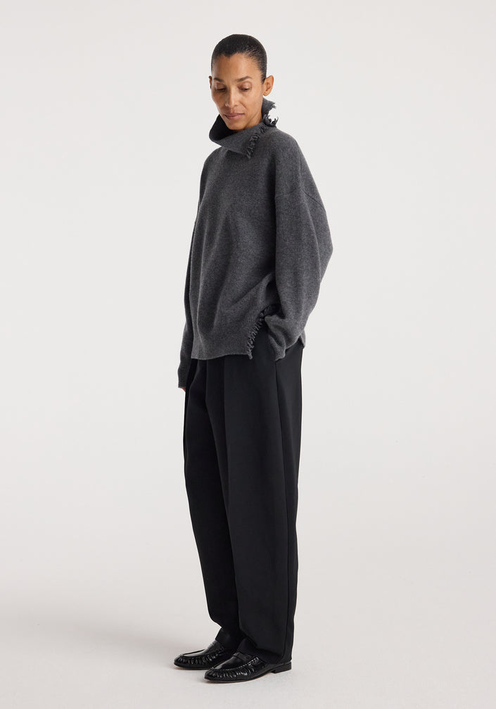 Model wearing a loose-fitting dark grey melange Turtleneck with raw details paired with wide-leg black trousers and black shoes against a plain white background.