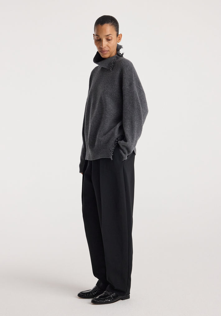Woman wearing a loose-fitting Turtleneck with raw details | dark grey melange, paired with wide-leg black trousers and black shoes, standing against a plain white background.