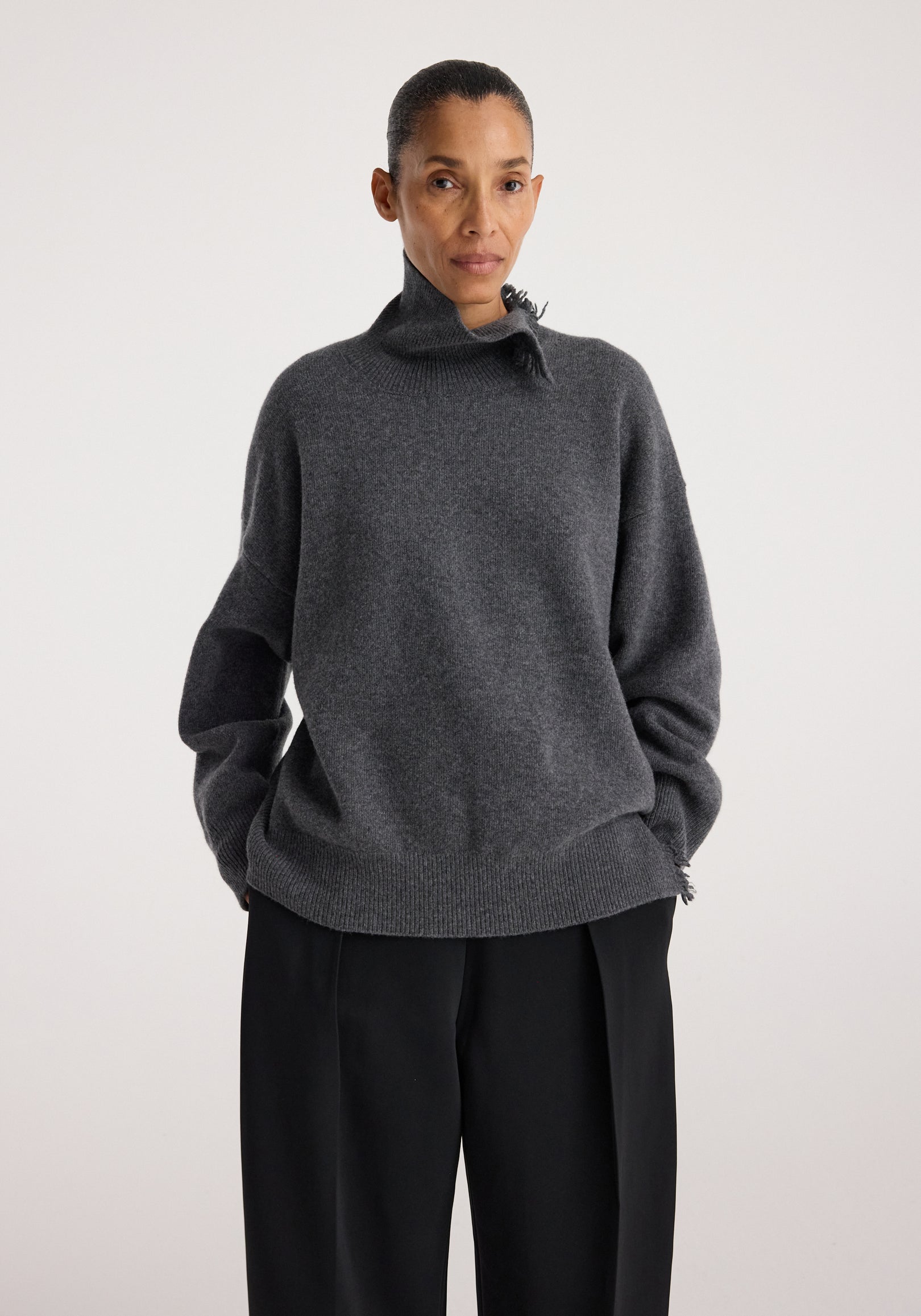 Woman wearing a dark grey melange Turtleneck with raw details, featuring a high neck and relaxed fit paired with black pants against a plain background.