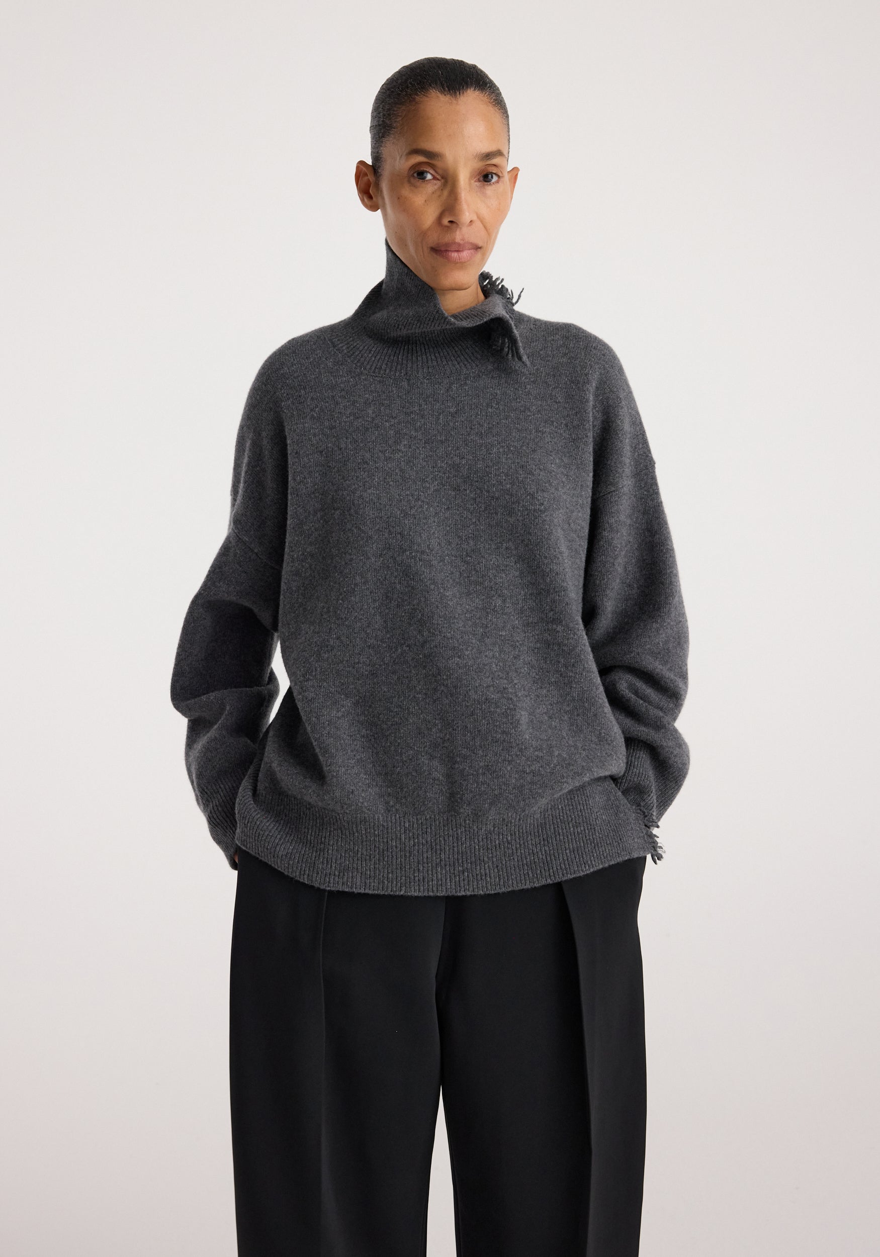 Woman wearing a dark grey melange Turtleneck with raw details, featuring a high neck and relaxed fit paired with black pants against a plain background.