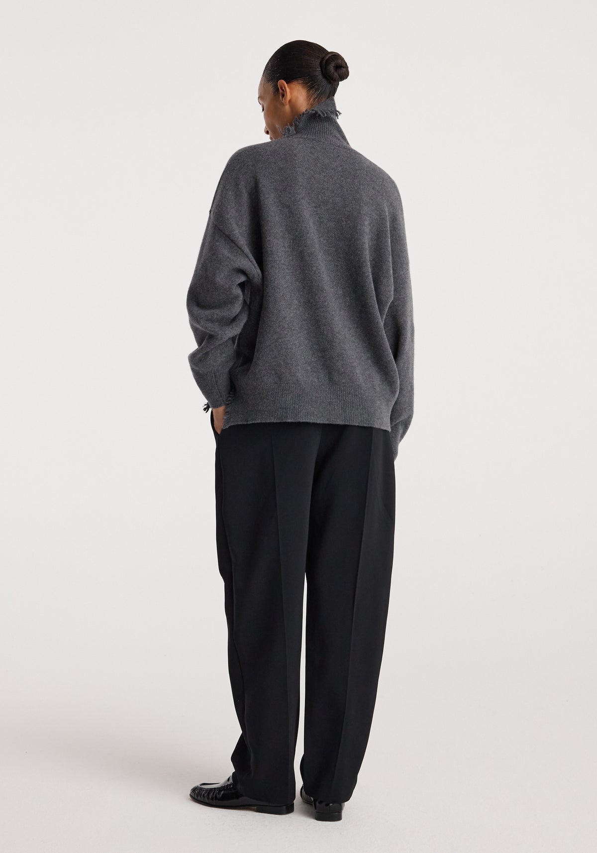 Woman wearing a dark grey melange turtleneck with raw details, paired with loose black pants and black shoes, standing against a plain light background.