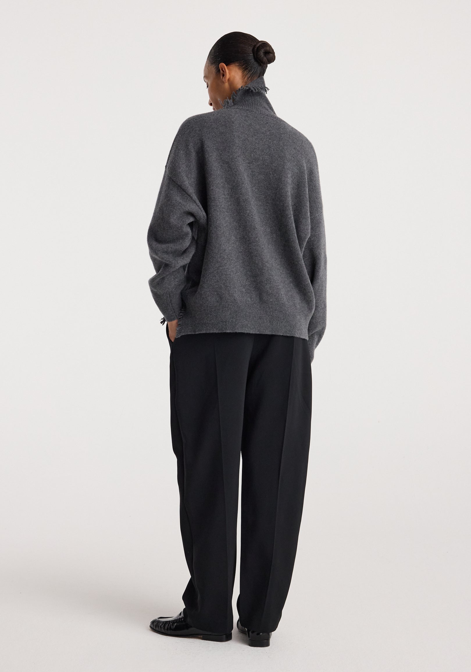 Woman wearing a dark grey melange turtleneck with raw details, paired with loose black pants and black shoes, standing against a plain light background.