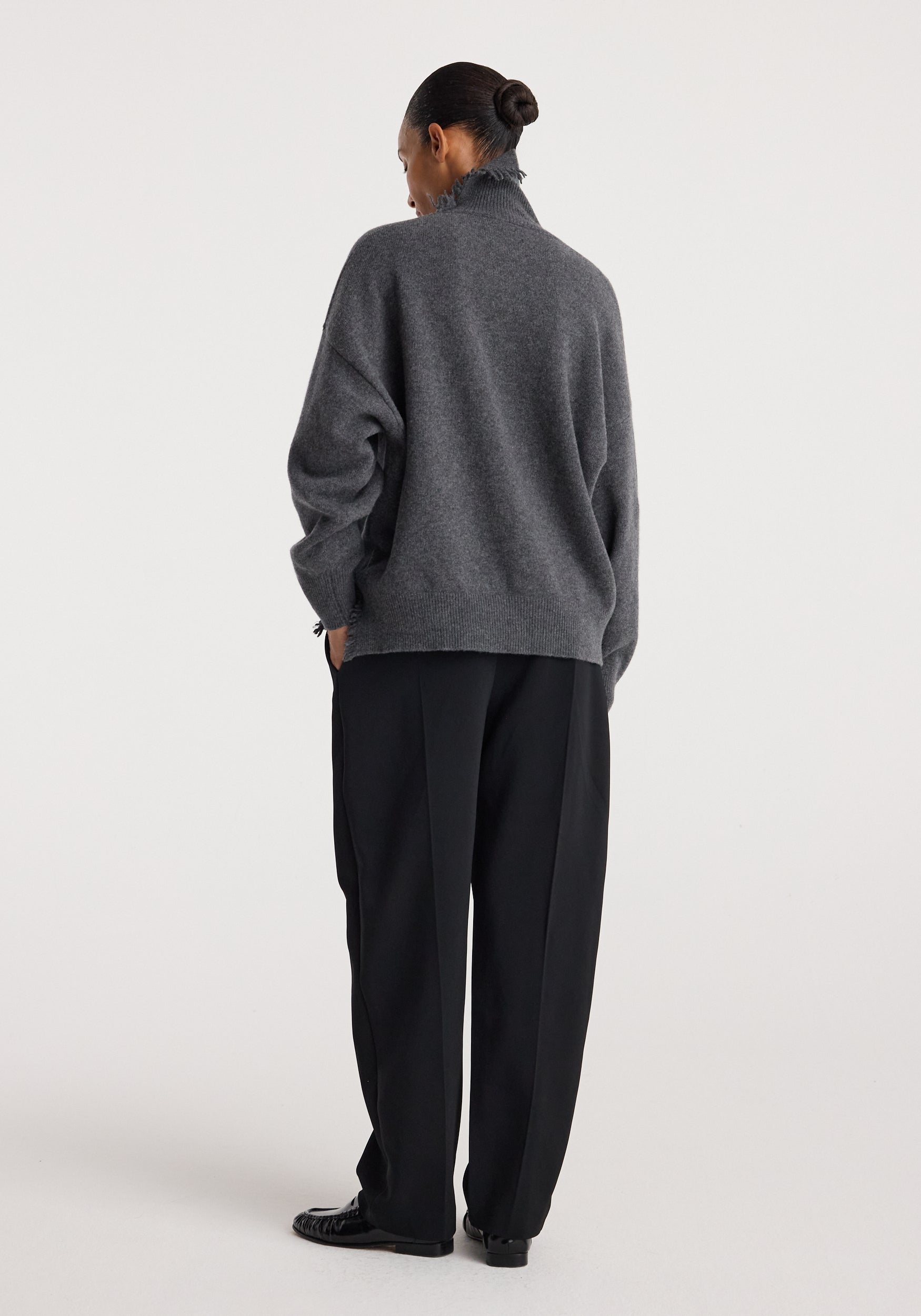 Woman wearing a dark grey melange turtleneck with raw details, paired with loose black pants and black shoes, standing against a plain light background.