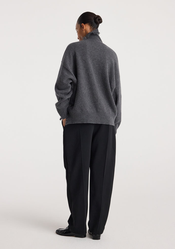 Woman wearing a dark grey melange turtleneck with raw details, paired with loose black pants and black shoes, standing against a plain light background.