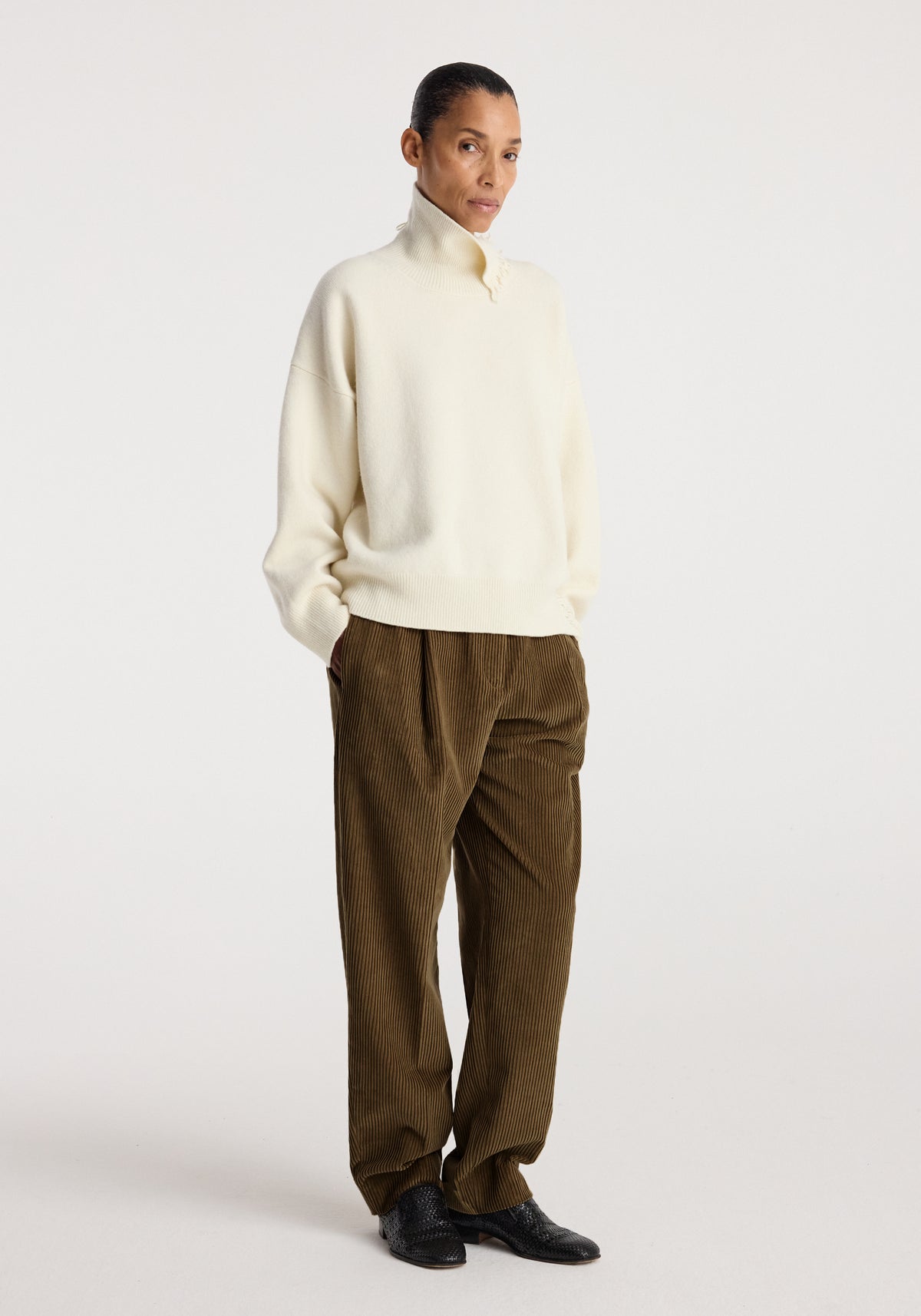 Woman wearing an off-white turtleneck with raw details paired with loose brown corduroy pants and black shoes, standing against a plain background.