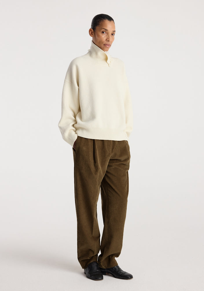 Woman wearing an off-white turtleneck with raw details paired with loose brown corduroy pants and black shoes, standing against a plain background.