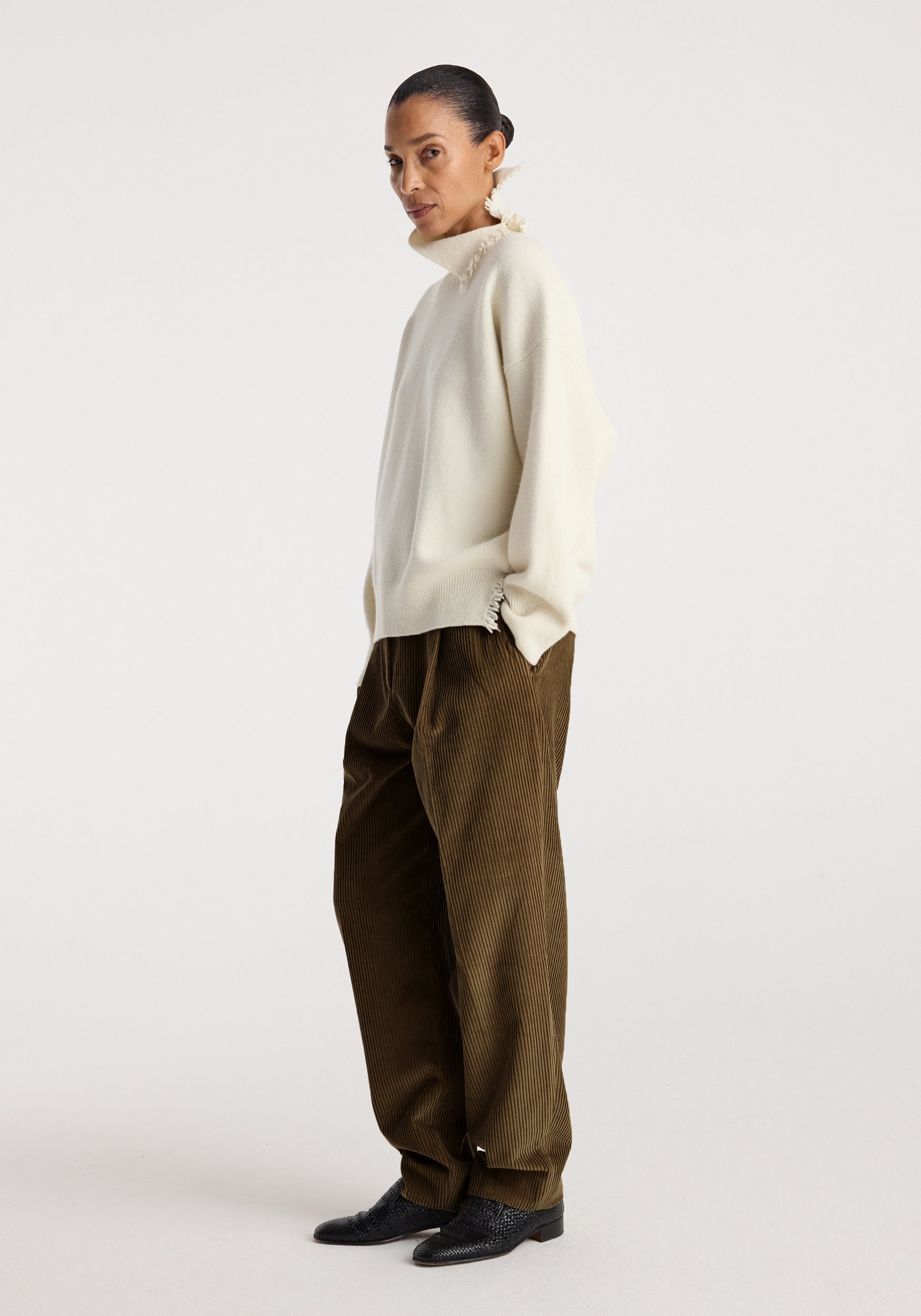Woman wearing a Turtleneck with raw details | off white styled with brown wide-leg corduroy pants and black textured shoes, standing against a plain white background.