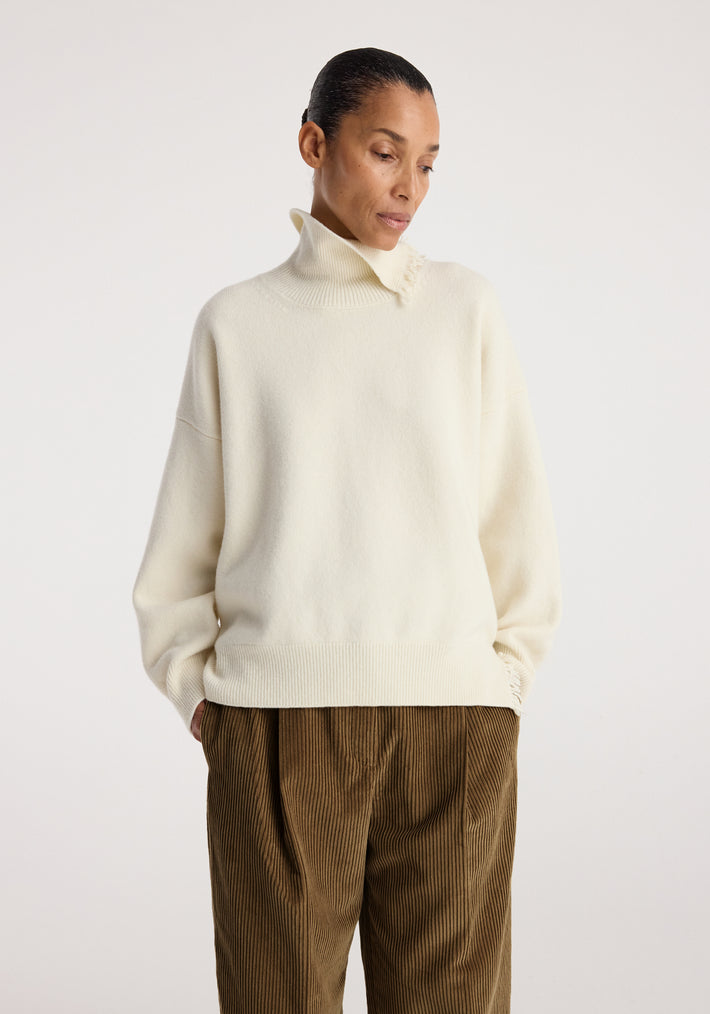 Woman wearing a cozy off white Turtleneck with raw details, featuring a high collar and relaxed fit, paired with brown corduroy pants against a plain light background.