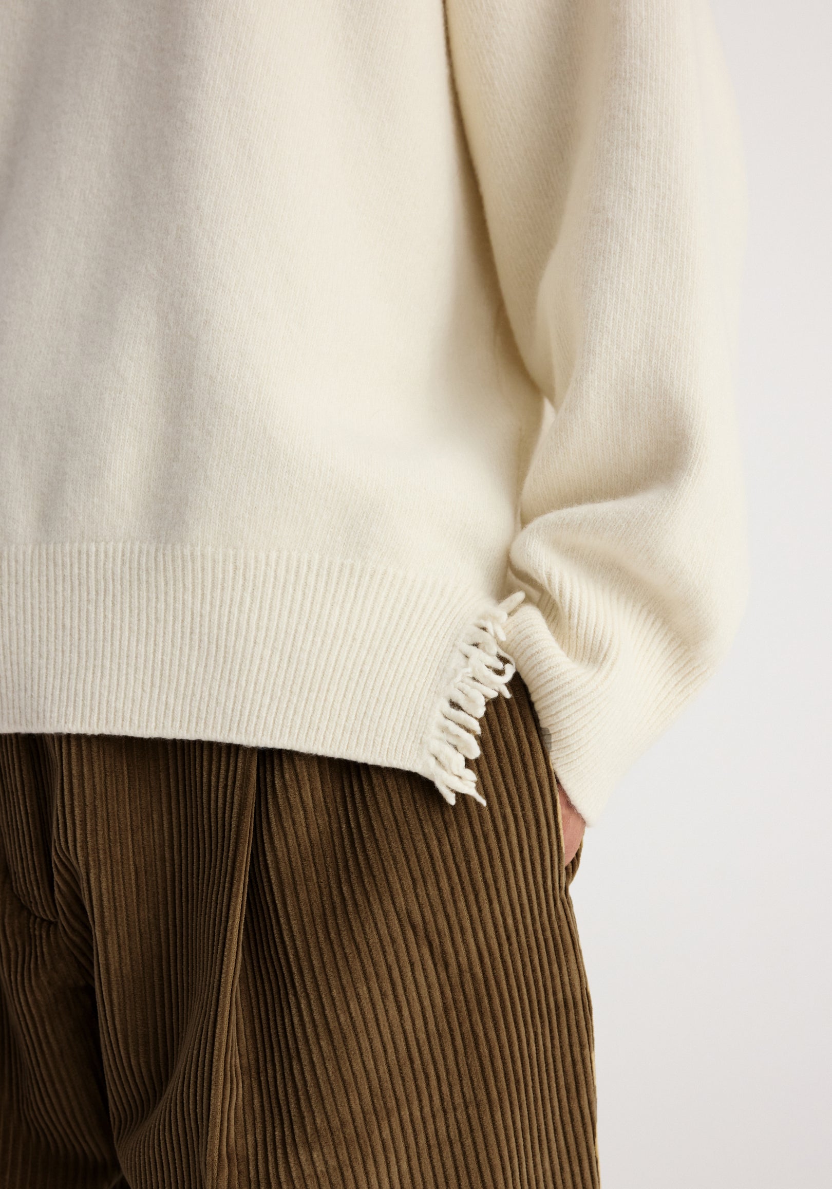 Close-up of a person wearing a Turtleneck with raw details | off white, highlighting the textured knit fabric and frayed hem detail paired with brown corduroy pants.