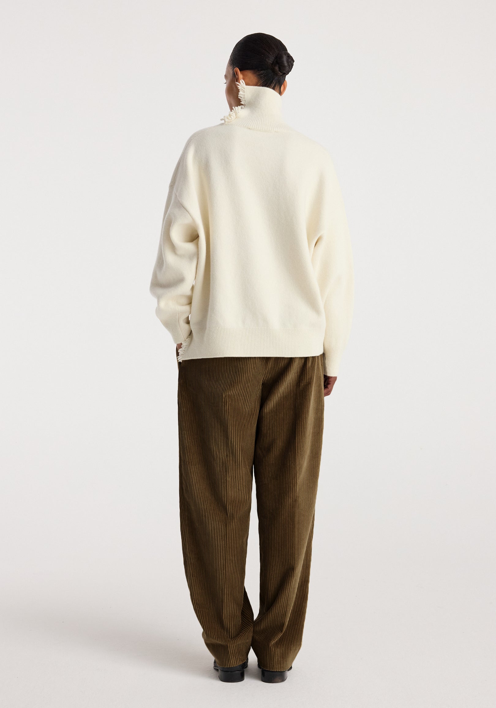 Woman wearing an off white turtleneck with raw details, styled with loose brown corduroy pants, standing facing away against a plain light background.