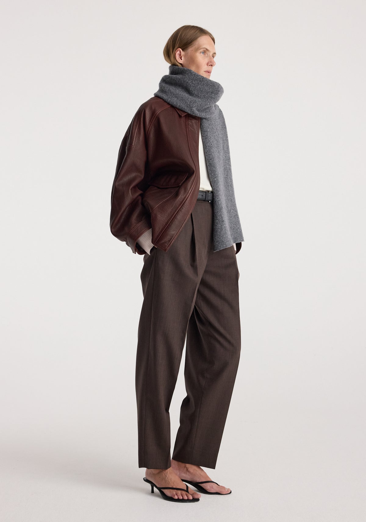 Woman wearing a Knitted mouliné scarf | anthracite melange wrapped around her neck paired with a brown leather jacket, dark brown trousers, and black heeled sandals.