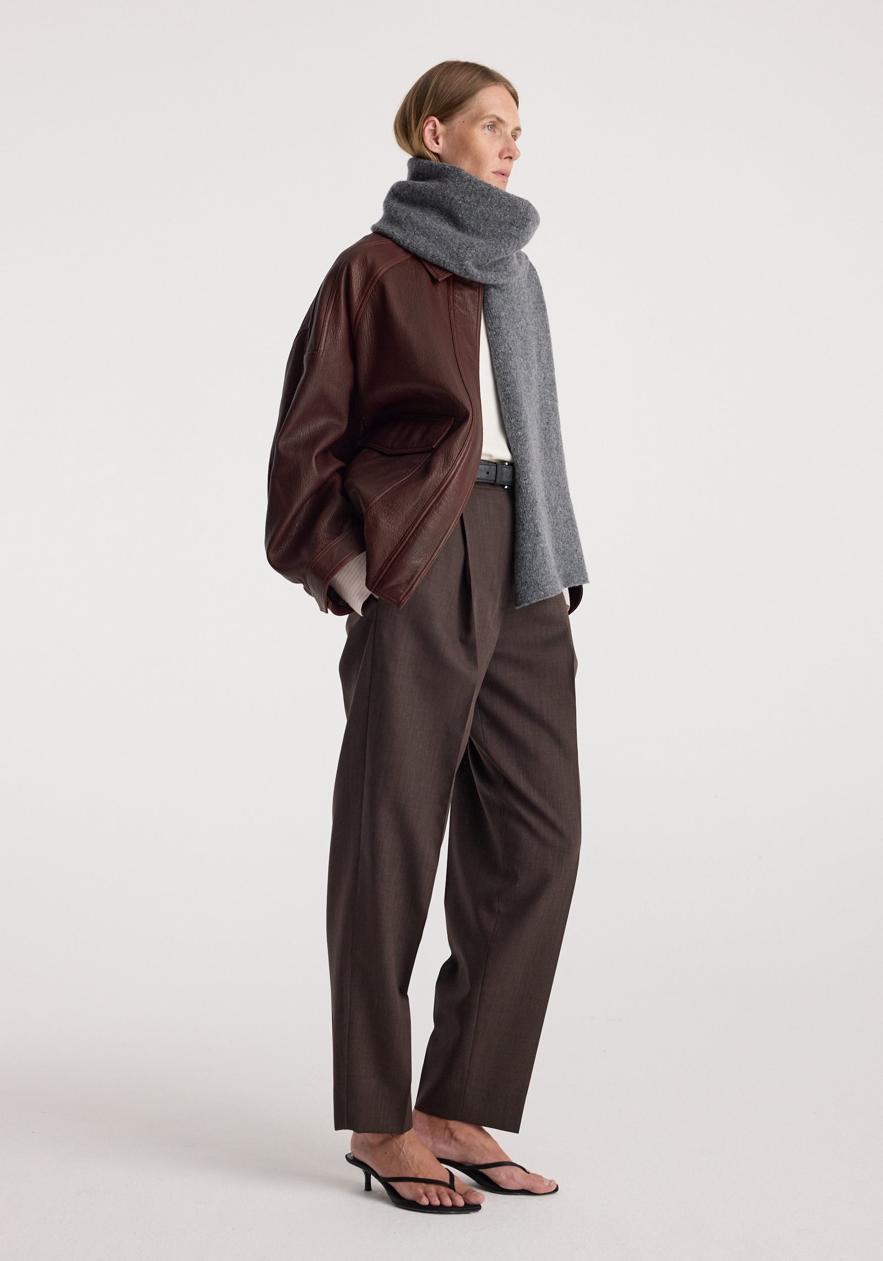 Woman wearing a Knitted mouliné scarf | anthracite melange wrapped around her neck paired with a brown leather jacket, dark brown trousers, and black heeled sandals.