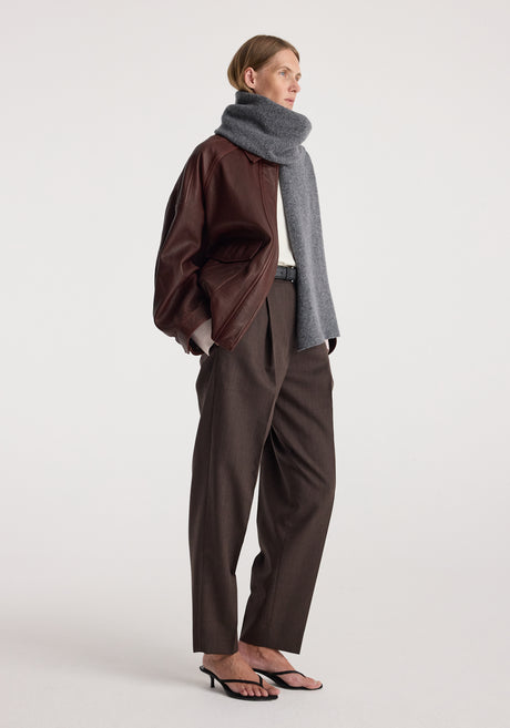 Woman wearing a Knitted mouliné scarf | anthracite melange wrapped around her neck paired with a brown leather jacket, dark brown trousers, and black heeled sandals.